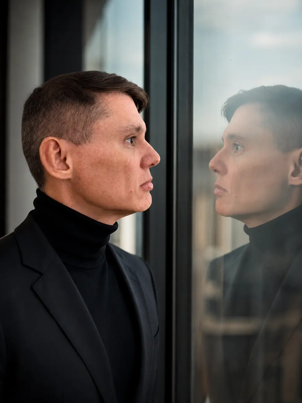 Aldo Batista de perfil diante de uma janela, com o reflexo ao lado